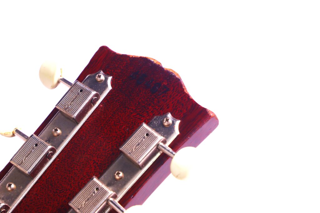 Nahaufnahme Der Rückseite Einer Gibson J-45 1962 Akustik Gitarre Mit Metallstimmwirbeln Und Einer Rötlichen Holzoberfläche Vor Einem Strahlend Weißen Hintergrund.