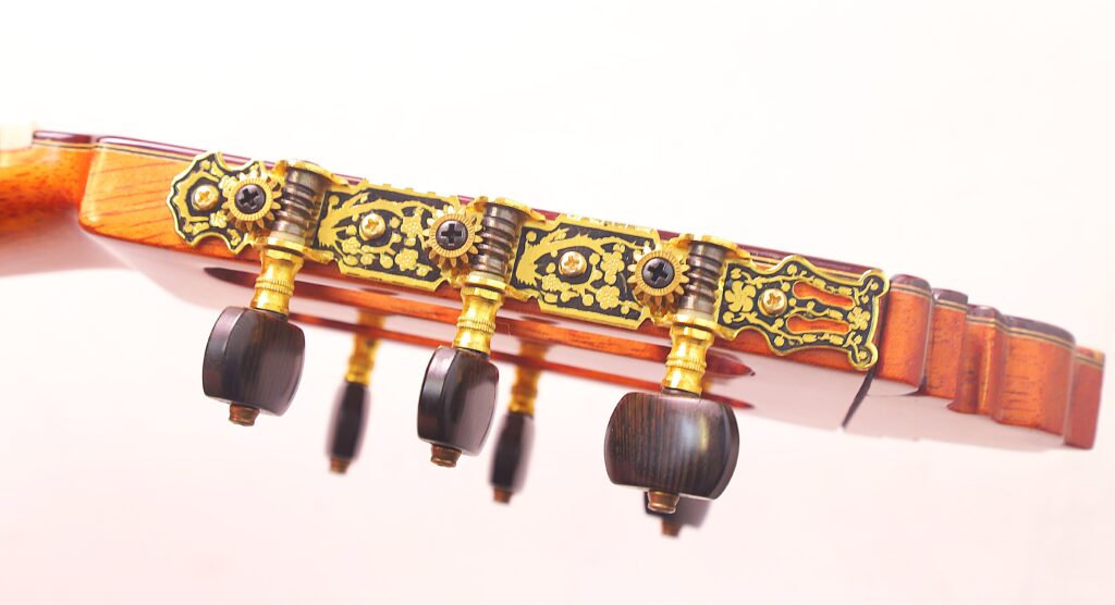 Nahaufnahme Von Drei Verzierten Stimmwirbeln An Der Pedro Fernández Luque 2020 Blanca Flamenco-Gitarre, Die Goldene Und Schwarze Dekorative Metallplatten Mit Dunklen Holzwirbeln Vor Einem Hellen Hintergrund Zeigt.