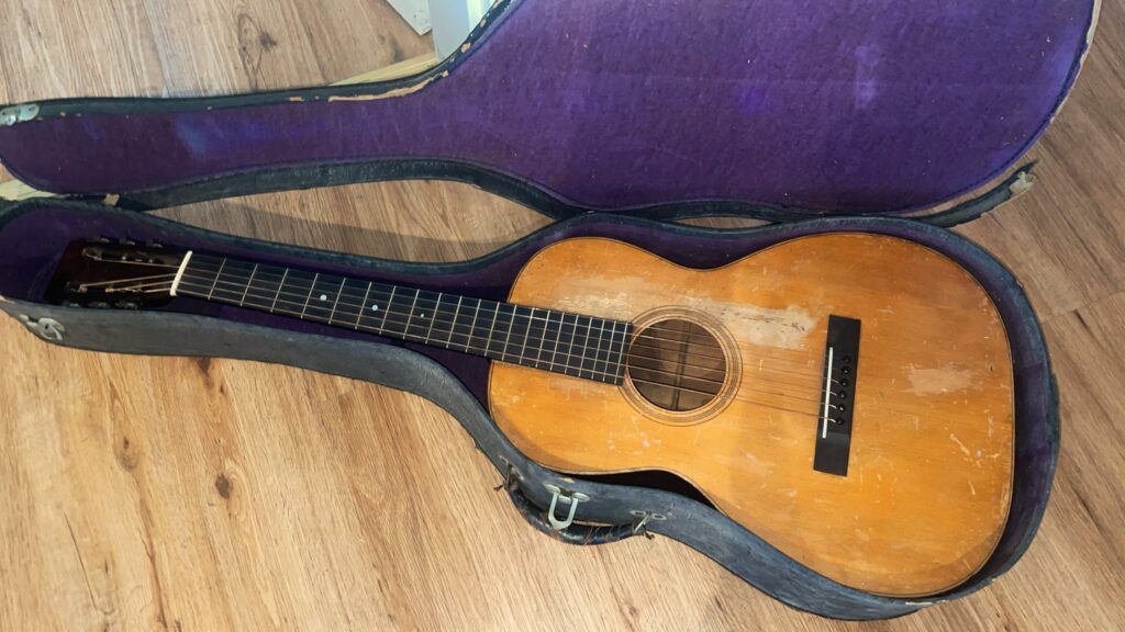 Eine Martin 0-18 1926 Akustik-Gitarre Mit Holzoberfläche Liegt In Einem Offenen, Mit Lila Stoff Ausgekleideten Vintage-Hartschalenkoffer Auf Einem Holzboden.