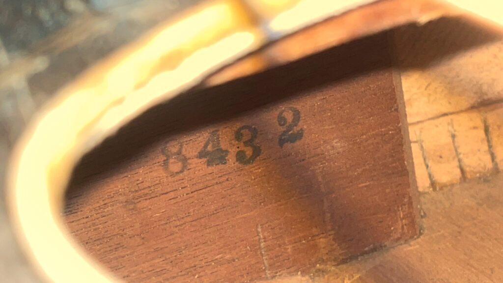 Eine Nahaufnahme Des Innenraums Der Gibson L-1 1926 Akustik Gitarre Zeigt Die Mit Schwarzer Tinte In Das Holz Gestempelte Nummer 8432, Die Durch Die Warme Beleuchtung Hervorgehoben Wird.