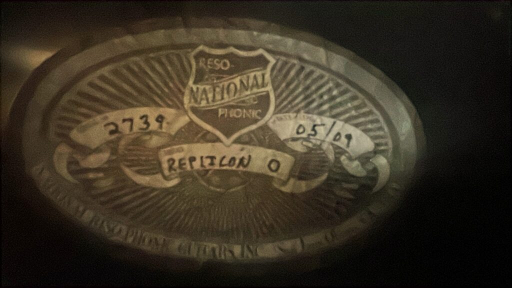 Ein Ovales Metallabzeichen Auf Der National Reso-Phonic Style O 2009 Akustik Gitarre Zeigt Ein Schild Mit &Quot;National&Quot; Und &Quot;Reso Phonic&Quot;, Handschriftliche Angaben (2739, 05/09, Repiton O) Und Ein Von Der Mitte Ausstrahlendes Sonnenschliffmuster.