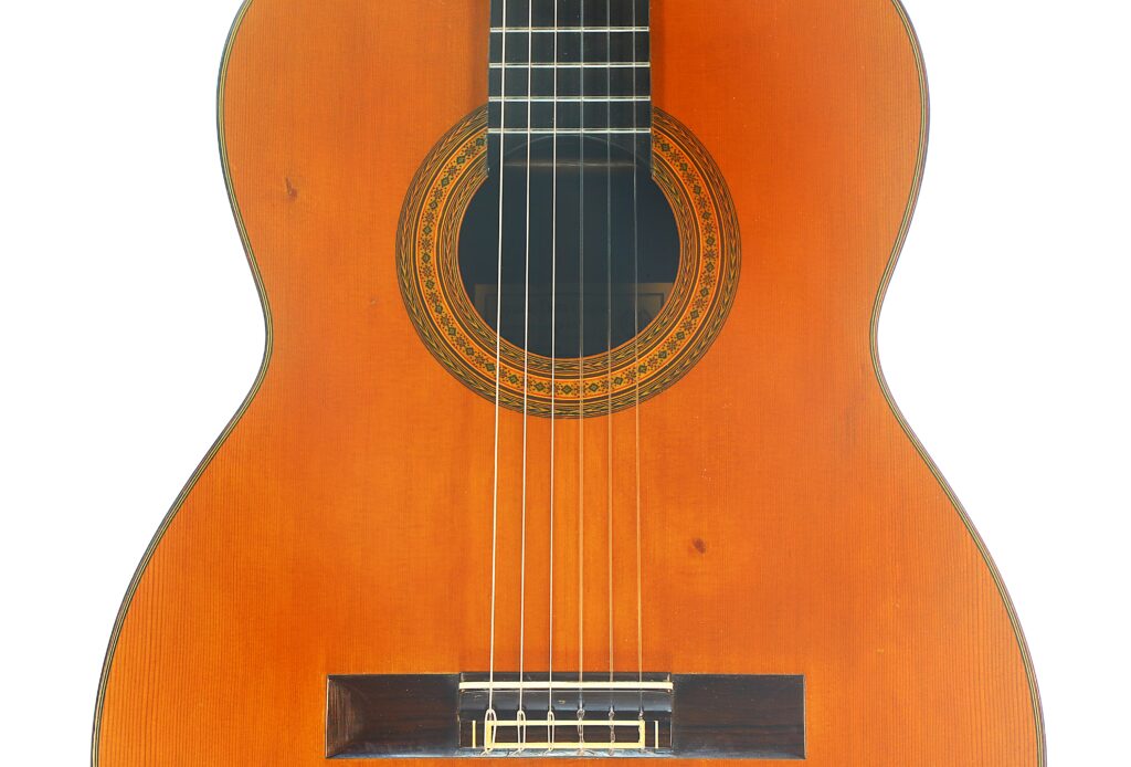 Nahaufnahme der „Ignacio Fleta 1992 Klassische Gitarre“ mit sechs Saiten, einer aufwendig verzierten Rosette, die das Schallloch umgibt, sowie Teilen des Holzkorpus und des Stegs. Nahaufnahme der „Ignacio Fleta 1992 Klassische Gitarre“ mit sechs Saiten, einer aufwendig verzierten Rosette, die das Schallloch umgibt, sowie Teilen des Holzkorpus und des Stegs.