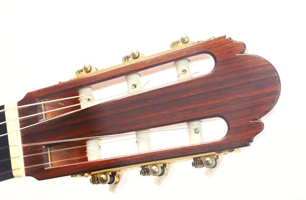 Eine Detailansicht des Kopfes der „Ignacio Fleta 1992 Klassische Gitarre“ mit Holzoberfläche, sechs Saiten und weißen Stimmwirbeln, fotografiert vor einem schlichten weißen Hintergrund. Eine Detailansicht des Kopfes der „Ignacio Fleta 1992 Klassische Gitarre“ mit Holzoberfläche, sechs Saiten und weißen Stimmwirbeln, fotografiert vor einem schlichten weißen Hintergrund.