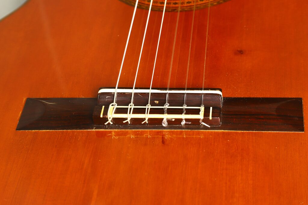 Eine Nahaufnahme der „Ignacio Fleta 1992 Klassische Gitarre“ hebt den wunderschön gearbeiteten Steg und die Saitenhalter an ihrem sattebraunen Holzkorpus hervor, wobei der Fokus auf dem unteren Teil des Instruments liegt. Eine Nahaufnahme der „Ignacio Fleta 1992 Klassische Gitarre“ hebt den wunderschön gearbeiteten Steg und die Saitenhalter an ihrem sattebraunen Holzkorpus hervor, wobei der Fokus auf dem unteren Teil des Instruments liegt.