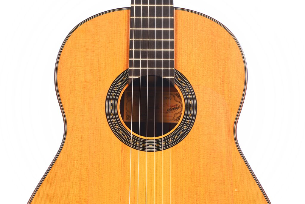 Nahaufnahme der Antonio Morales Nogués 2020 Negra Flamenco-Gitarre. Hervorzuheben sind die sechs Saiten, der hölzerne Resonanzboden, das dekorative runde Schallloch und ein Teil des Griffbretts vor einem weißen Hintergrund. Nahaufnahme der Antonio Morales Nogués 2020 Negra Flamenco-Gitarre. Hervorzuheben sind die sechs Saiten, der hölzerne Resonanzboden, das dekorative runde Schallloch und ein Teil des Griffbretts vor einem weißen Hintergrund.