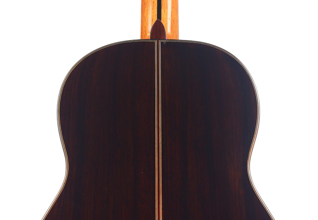 Das Bild zeigt die Rückseite einer Antonio Morales Nogués 2020 Negra Flamenco-Gitarre, gefertigt aus dunklem, poliertem Holz mit einer hellen dekorativen Mittelnaht und Randeinfassung. Der Hals ist teilweise sichtbar an der Spitze vor einem weißen Hintergrund. Das Bild zeigt die Rückseite einer Antonio Morales Nogués 2020 Negra Flamenco-Gitarre, gefertigt aus dunklem, poliertem Holz mit einer hellen dekorativen Mittelnaht und Randeinfassung. Der Hals ist teilweise sichtbar an der Spitze vor einem weißen Hintergrund.