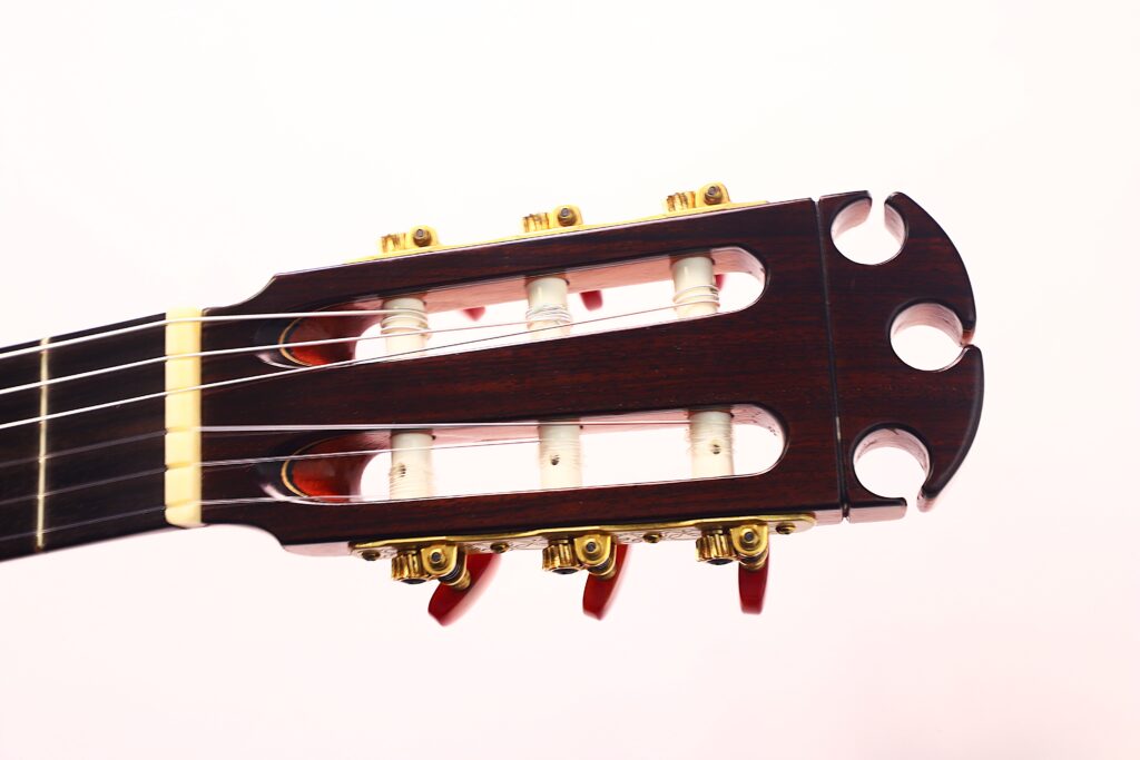 Nahaufnahme der Kopfplatte einer Antonio Morales Nogués 2020 Negra Flamenco-Gitarre mit goldenen Stimmwirbeln und drei Nylonsaiten, die vor einem schlichten hellen Hintergrund zu sehen sind. Nahaufnahme der Kopfplatte einer Antonio Morales Nogués 2020 Negra Flamenco-Gitarre mit goldenen Stimmwirbeln und drei Nylonsaiten, die vor einem schlichten hellen Hintergrund zu sehen sind.