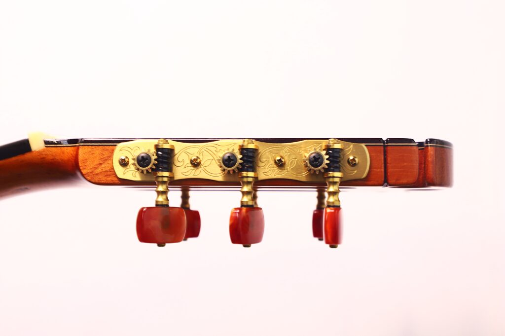Nahaufnahme der Stimmwirbel an der Kopfplatte einer Antonio Morales Nogués 2020 Negra Flamenco-Gitarre mit goldener Hardware und roten Stimmknöpfen vor einem schlichten, hellen Hintergrund. Nahaufnahme der Stimmwirbel an der Kopfplatte einer Antonio Morales Nogués 2020 Negra Flamenco-Gitarre mit goldener Hardware und roten Stimmknöpfen vor einem schlichten, hellen Hintergrund.