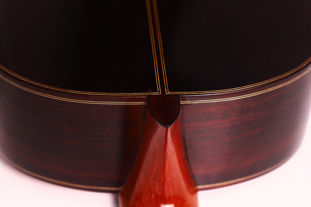 Nahaufnahme des Bodens und des Halses der Antonio Morales Nogués 2020 Negra Flamenco-Gitarre. Sie zeigt die polierte Holzoberfläche, die dekorativen Intarsien und die Beleuchtung, die die reichen dunkelbraunen und rötlichen Holztöne hervorhebt. Nahaufnahme des Bodens und des Halses der Antonio Morales Nogués 2020 Negra Flamenco-Gitarre. Sie zeigt die polierte Holzoberfläche, die dekorativen Intarsien und die Beleuchtung, die die reichen dunkelbraunen und rötlichen Holztöne hervorhebt.