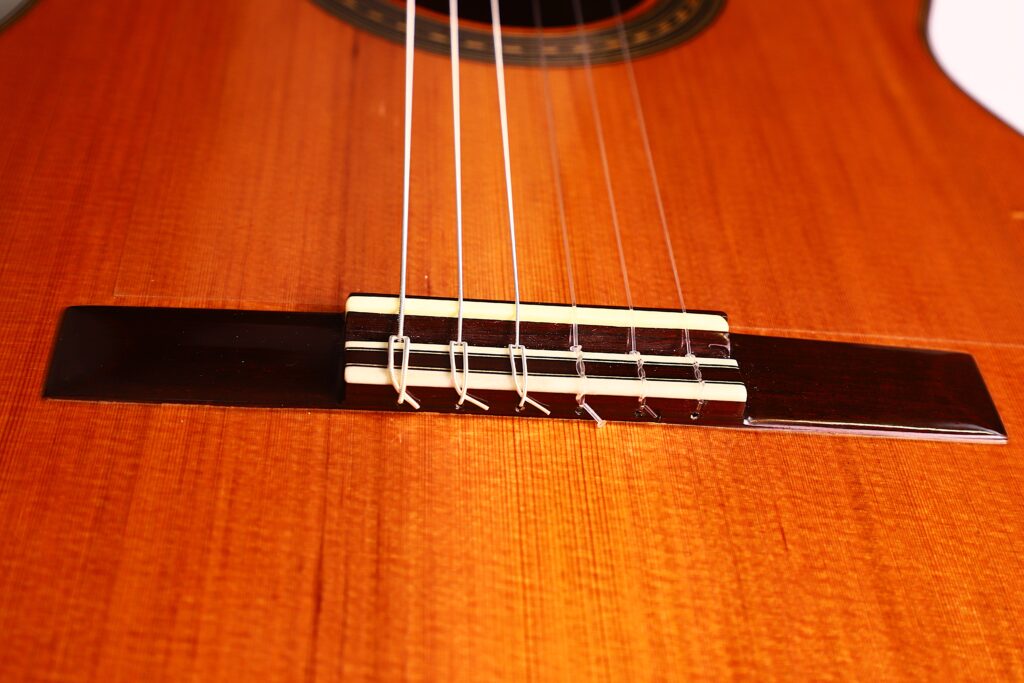 Nahaufnahme des Stegs der Antonio Morales Nogués 2020 Negra Flamenco Gitarre mit sechs Saiten, die die Holzmaserung und die detaillierte Handwerkskunst dieses klassischen Instruments hervorhebt. Nahaufnahme des Stegs der Antonio Morales Nogués 2020 Negra Flamenco Gitarre mit sechs Saiten, die die Holzmaserung und die detaillierte Handwerkskunst dieses klassischen Instruments hervorhebt.