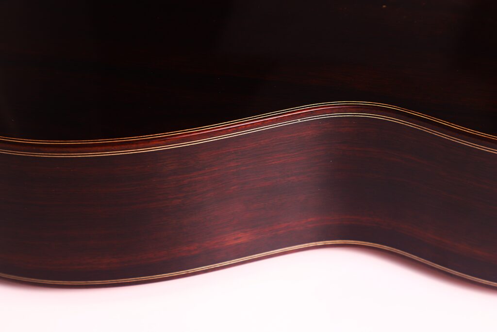 Nahaufnahme der dunklen Holzseite der Antonio Morales Nogués 2020 Negra Flamenco-Gitarre. Hervorzuheben sind die sanft geschwungene Form und die feinen Kantendetails, die sich von einer hellrosa Oberfläche abheben. Nahaufnahme der dunklen Holzseite der Antonio Morales Nogués 2020 Negra Flamenco-Gitarre. Hervorzuheben sind die sanft geschwungene Form und die feinen Kantendetails, die sich von einer hellrosa Oberfläche abheben.