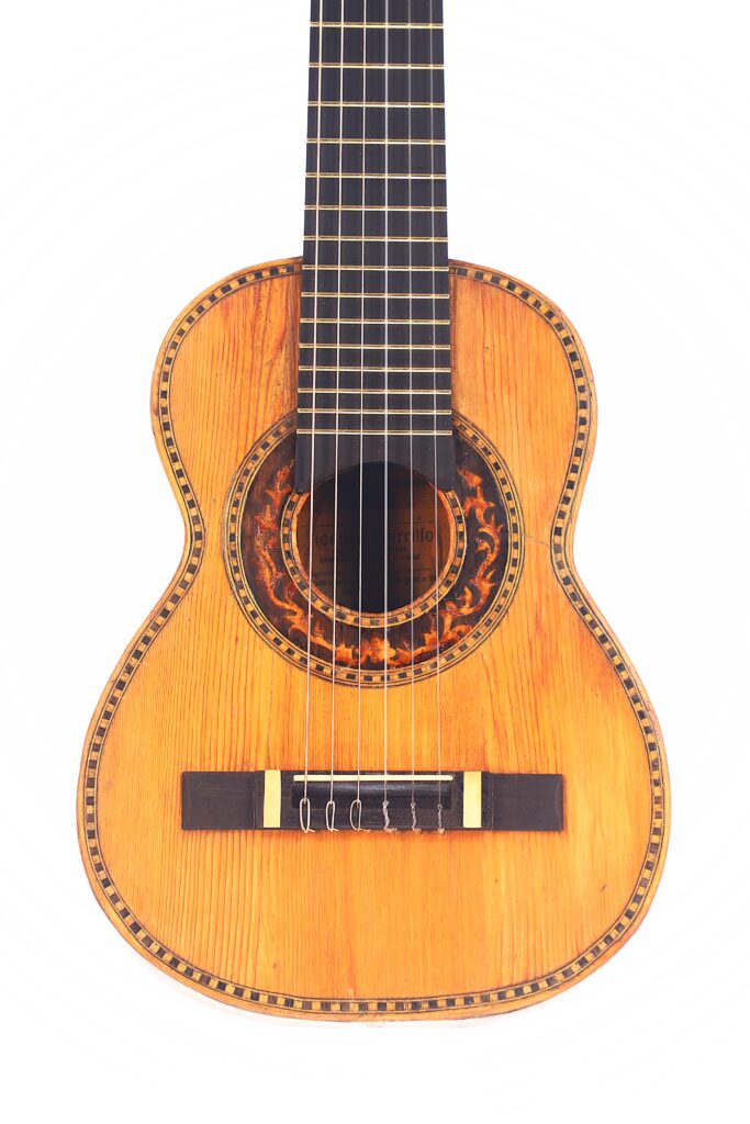 Die Vicente Carillo 1925 Terz Gitarre ist eine klassische Akustikgitarre mit einer natürlichen Holzoberfläche, einer verzierten Rosette um das Schallloch, sechs Saiten und einem sichtbaren Steg, fotografiert vor einem weißen Hintergrund. Die Vicente Carillo 1925 Terz Gitarre ist eine klassische Akustikgitarre mit einer natürlichen Holzoberfläche, einer verzierten Rosette um das Schallloch, sechs Saiten und einem sichtbaren Steg, fotografiert vor einem weißen Hintergrund.