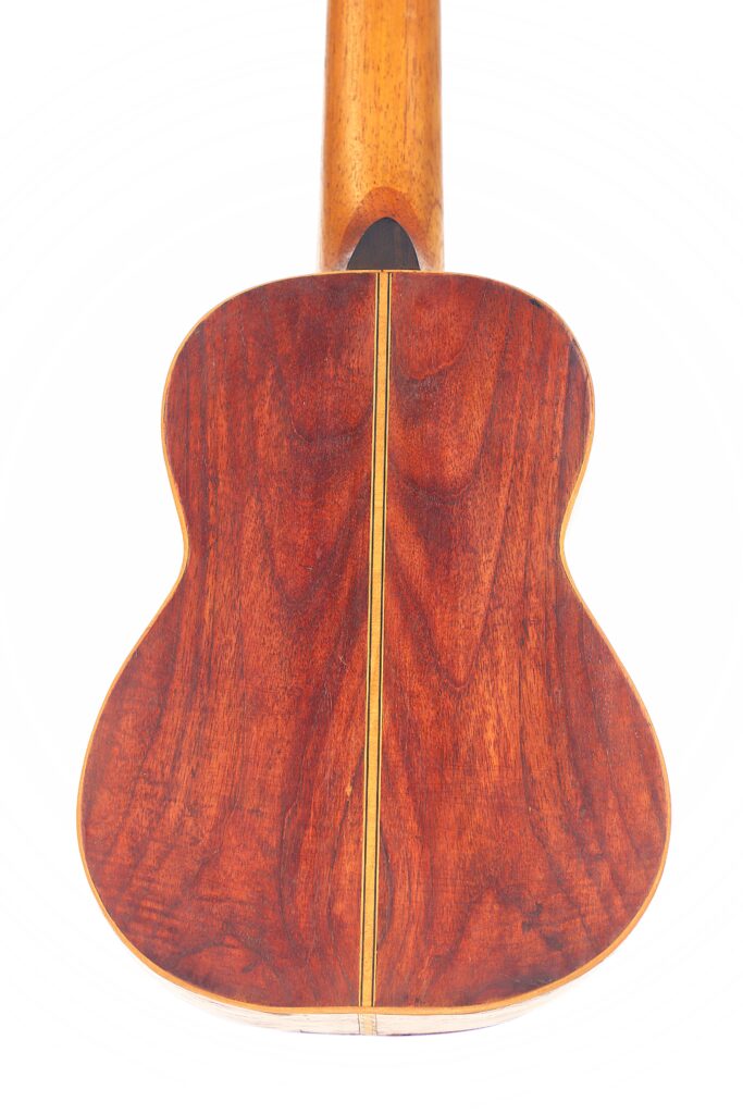 Eine Nahaufnahme der Rückseite der Vicente Carillo 1925 Terz Gitarre hebt die rötlich-braune Holzmaserung und die zentrale vertikale Einlage vor weißem Hintergrund hervor. Eine Nahaufnahme der Rückseite der Vicente Carillo 1925 Terz Gitarre hebt die rötlich-braune Holzmaserung und die zentrale vertikale Einlage vor weißem Hintergrund hervor.