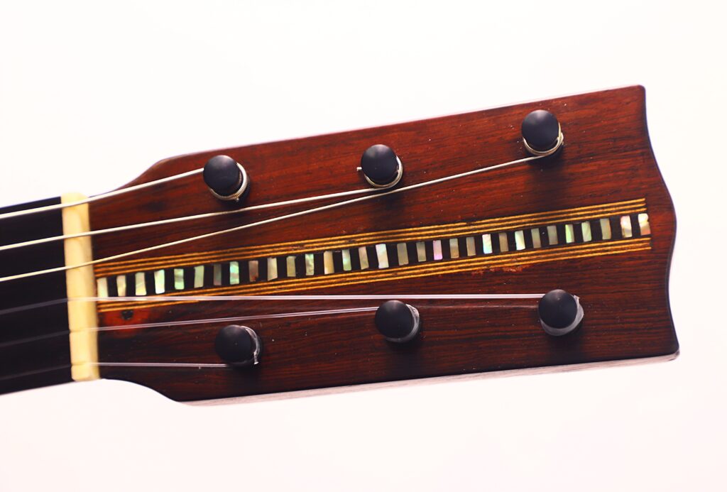 Nahaufnahme der hölzernen Kopfplatte der Franciso Pau 1890 Klassische Gitarre mit sechs Stiftwirbeln, dekorativer Einlage und angebrachten Saiten, vor einem schlichten weißen Hintergrund. Nahaufnahme der hölzernen Kopfplatte der Franciso Pau 1890 Klassische Gitarre mit sechs Stiftwirbeln, dekorativer Einlage und angebrachten Saiten, vor einem schlichten weißen Hintergrund.