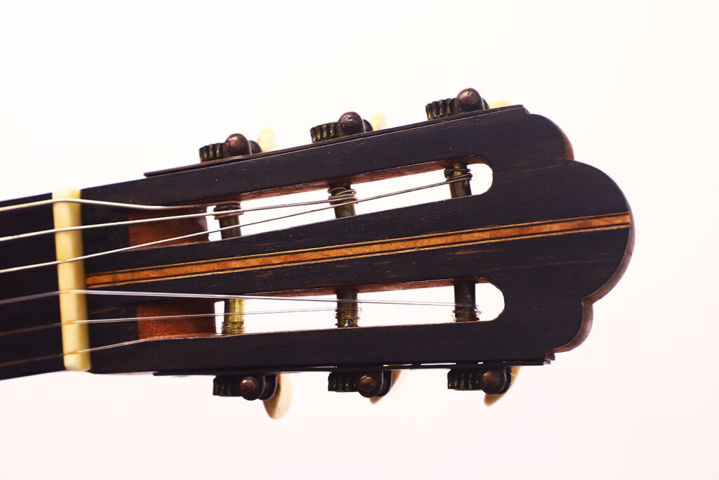 Nahaufnahme der Kopfplatte der Vicente Carillo 1925 Terz Gitarre mit Stimmwirbeln, Nylonsaiten und einer Holzoberfläche vor einem schlichten weißen Hintergrund. Nahaufnahme der Kopfplatte der Vicente Carillo 1925 Terz Gitarre mit Stimmwirbeln, Nylonsaiten und einer Holzoberfläche vor einem schlichten weißen Hintergrund.
