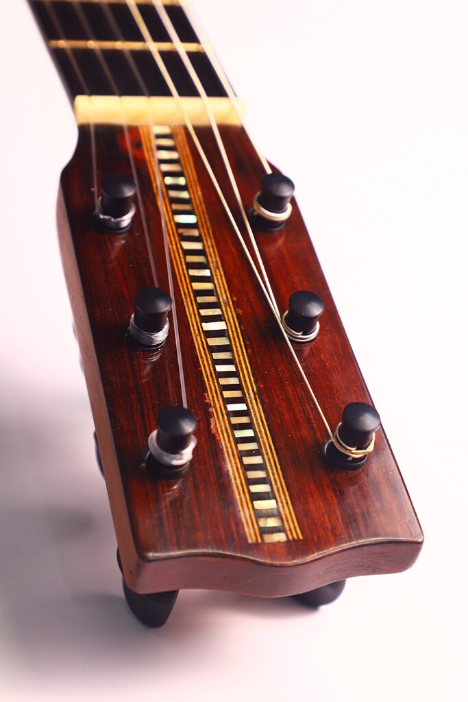 Nahaufnahme der hölzernen Kopfplatte der Franciso Pau 1890 Klassische Gitarre mit schwarzen Stimmwirbeln, Gold- und Weißeinlage in der Mitte und Gitarrensaiten, die bis zum Hals reichen, fotografiert vor einem hellen Hintergrund. Nahaufnahme der hölzernen Kopfplatte der Franciso Pau 1890 Klassische Gitarre mit schwarzen Stimmwirbeln, Gold- und Weißeinlage in der Mitte und Gitarrensaiten, die bis zum Hals reichen, fotografiert vor einem hellen Hintergrund.
