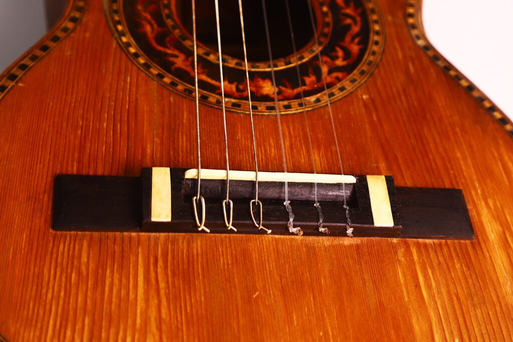 Nahaufnahme der Vicente Carillo 1925 Terz Gitarre, die den hölzernen Korpus, den dunklen Steg mit sechs Saiten und die detaillierte Einlage um das Schallloch hervorhebt. Nahaufnahme der Vicente Carillo 1925 Terz Gitarre, die den hölzernen Korpus, den dunklen Steg mit sechs Saiten und die detaillierte Einlage um das Schallloch hervorhebt.