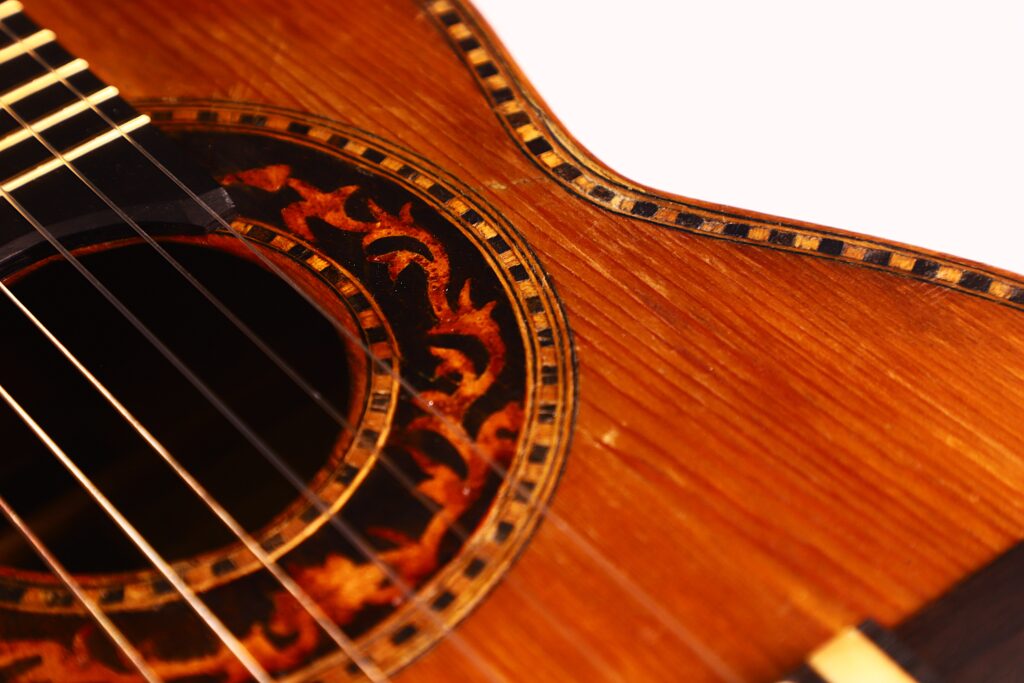 Nahaufnahme der Vicente Carillo 1925 Terz Gitarre, die den hölzernen Korpus, die verzierte Rosette um das Schallloch, die Saiten und einen Teil des Griffbretts vor einem hellen Hintergrund zeigt. Nahaufnahme der Vicente Carillo 1925 Terz Gitarre, die den hölzernen Korpus, die verzierte Rosette um das Schallloch, die Saiten und einen Teil des Griffbretts vor einem hellen Hintergrund zeigt.