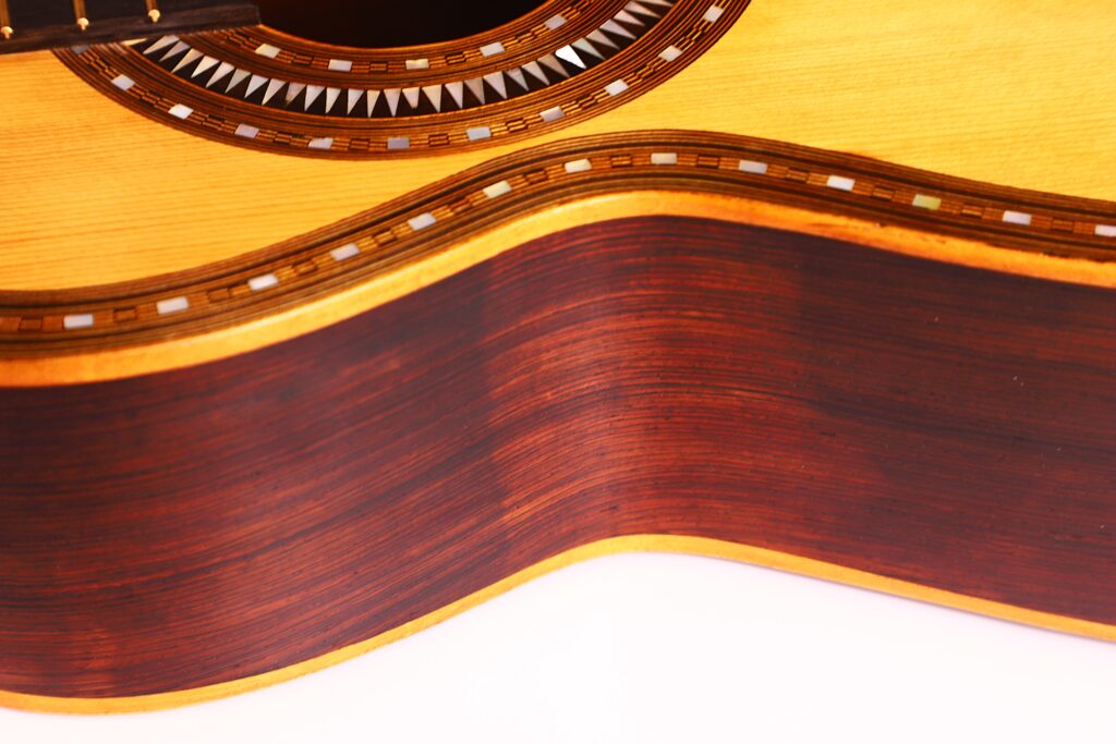 Nahaufnahme der Franciso Pau 1890 Klassische Gitarre mit ihrem Holzkorpus, der dekorativen Einlage um das Schallloch und der reichhaltigen, gewellten Holzmaserung auf der Seite vor weißem Hintergrund. Nahaufnahme der Franciso Pau 1890 Klassische Gitarre mit ihrem Holzkorpus, der dekorativen Einlage um das Schallloch und der reichhaltigen, gewellten Holzmaserung auf der Seite vor weißem Hintergrund.