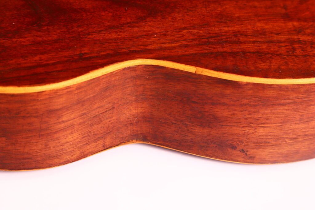 Nahaufnahme der Seite der Vicente Carillo 1925 Terz Gitarre. Hervorzuheben sind die glatte braune Oberfläche, die helleren Zierleisten und die eleganten Kurven vor einem weißen Hintergrund. Nahaufnahme der Seite der Vicente Carillo 1925 Terz Gitarre. Hervorzuheben sind die glatte braune Oberfläche, die helleren Zierleisten und die eleganten Kurven vor einem weißen Hintergrund.