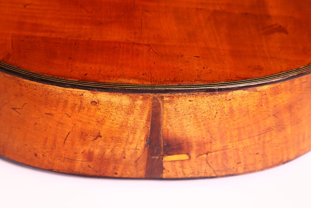 Nahaufnahme der Unterseite und des hinteren Randes einer C.F. Marchal 1820 Romantikgitarre, die ein warmes braunes Finish, sichtbare Holzmaserung, Kratzer und Gebrauchsspuren aufweist. Nahaufnahme der Unterseite und des hinteren Randes einer C.F. Marchal 1820 Romantikgitarre, die ein warmes braunes Finish, sichtbare Holzmaserung, Kratzer und Gebrauchsspuren aufweist.