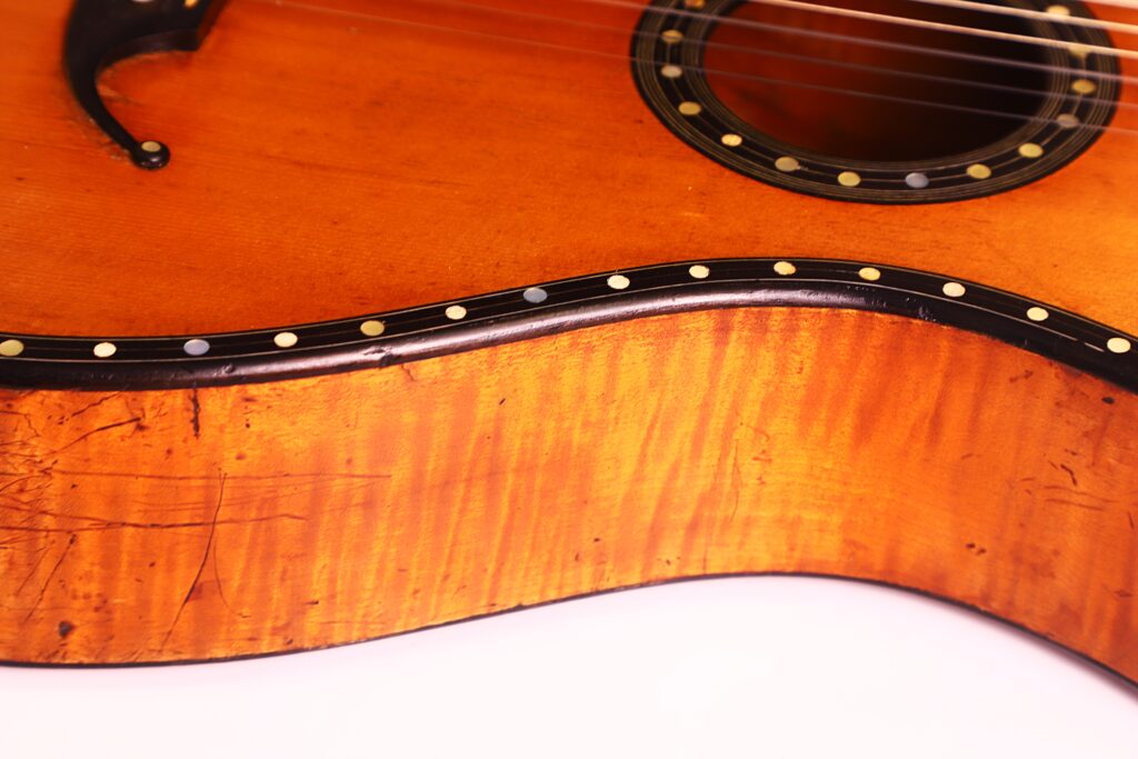 Nahaufnahme der C.F. Marchal 1820 Romantikgitarre, die die Holzmaserung, den dekorativen Schalllochring, die Saiten und eine Reihe kreisförmiger Intarsien entlang des Randes zeigt. Nahaufnahme der C.F. Marchal 1820 Romantikgitarre, die die Holzmaserung, den dekorativen Schalllochring, die Saiten und eine Reihe kreisförmiger Intarsien entlang des Randes zeigt.