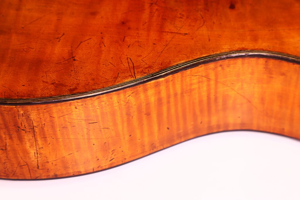 Nahaufnahme der Seite und der Rückseite einer C.F. Marchal 1820 Romantikgitarre, die eine warme, glänzende Oberfläche mit sichtbarer Holzmaserung und Kratzern auf weißem Hintergrund aufweist. Nahaufnahme der Seite und der Rückseite einer C.F. Marchal 1820 Romantikgitarre, die eine warme, glänzende Oberfläche mit sichtbarer Holzmaserung und Kratzern auf weißem Hintergrund aufweist.