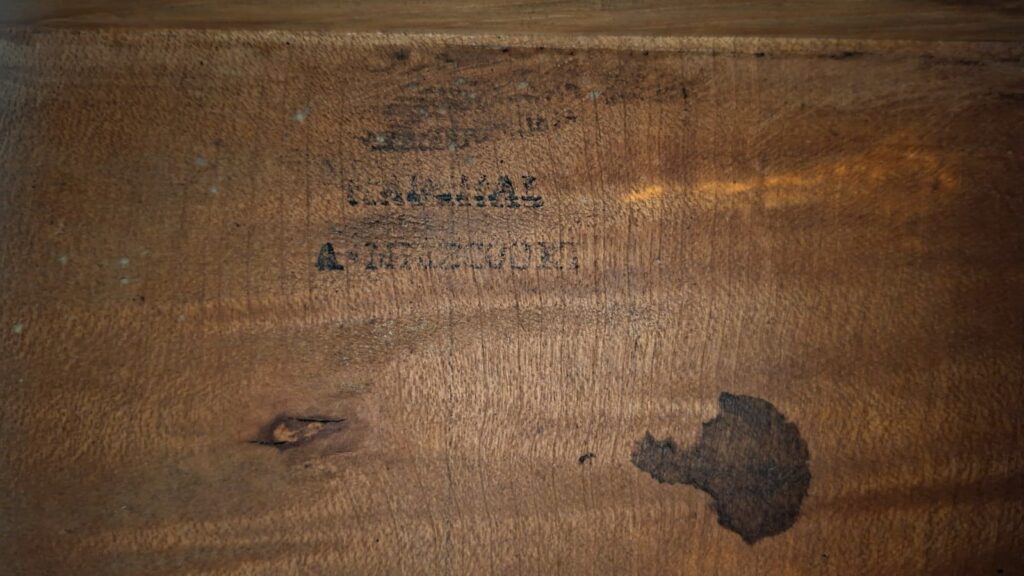 Eine Nahaufnahme der hölzernen Oberfläche der C.F. Marchal 1820 Romantikgitarre zeigt verblasste schwarze Schrift und Zahlen, von denen einige teilweise unleserlich sind, sowie einen dunklen, unregelmäßigen Fleck unten rechts. Eine Nahaufnahme der hölzernen Oberfläche der C.F. Marchal 1820 Romantikgitarre zeigt verblasste schwarze Schrift und Zahlen, von denen einige teilweise unleserlich sind, sowie einen dunklen, unregelmäßigen Fleck unten rechts.