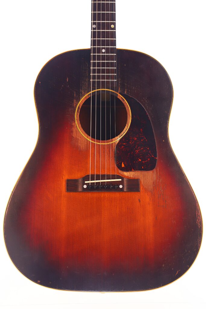 Eine Gibson J-45 1954 Akustische Gitarre mit Sunburst-Lackierung, sichtbarer Holzmaserung, einigen Gebrauchsspuren, einem abgerundeten Korpus, Schildpatt-Schlagbrett und weißen Punkt-Griffbretteinlagen. Eine Gibson J-45 1954 Akustische Gitarre mit Sunburst-Lackierung, sichtbarer Holzmaserung, einigen Gebrauchsspuren, einem abgerundeten Korpus, Schildpatt-Schlagbrett und weißen Punkt-Griffbretteinlagen.