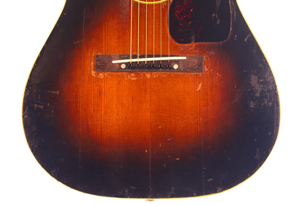 Die Nahaufnahme der Gibson LG-1 1953 Akustik Gitarre zeigt das Sunburst-Finish, die sichtbaren Kratzer, den Holzsteg, die sechs Saiten und das Schlagbrett mit Schildpattmuster. Die Nahaufnahme der Gibson LG-1 1953 Akustik Gitarre zeigt das Sunburst-Finish, die sichtbaren Kratzer, den Holzsteg, die sechs Saiten und das Schlagbrett mit Schildpattmuster.