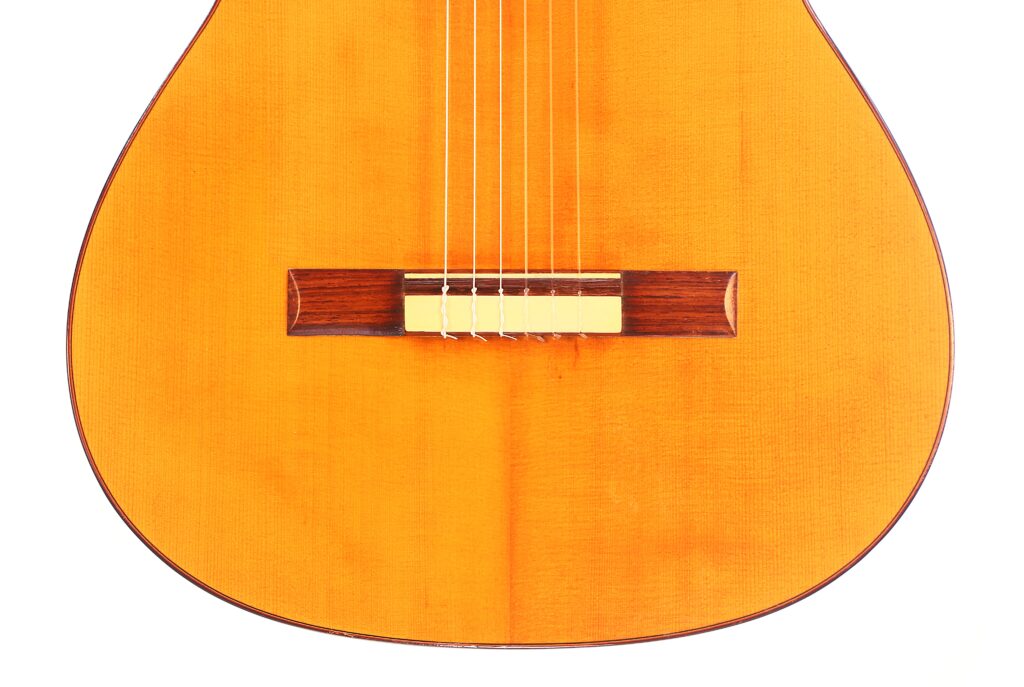 Nahaufnahme des Korpus der Marcelino López Nieto 1959 Flamenco Gitarre mit sechs Saiten, einem Holzsteg und einer warmen hellbraunen Lackierung. Das Bild zeigt die untere Hälfte der Vorderseite dieser klassischen Gitarre. Nahaufnahme des Korpus der Marcelino López Nieto 1959 Flamenco Gitarre mit sechs Saiten, einem Holzsteg und einer warmen hellbraunen Lackierung. Das Bild zeigt die untere Hälfte der Vorderseite dieser klassischen Gitarre.