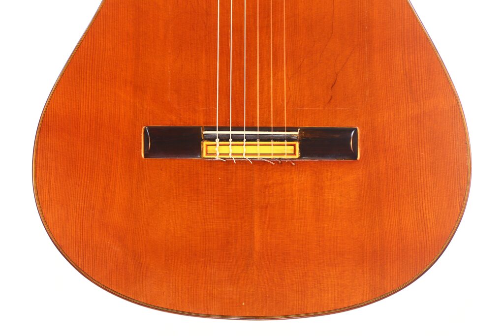 Nahaufnahme des Korpus der Hermanos Conde 1971 Faustino Conde Negra Flamenco-Gitarre. Hervorzuheben sind die Saiten, der Holzsteg und die warme braune Holzmaserung. Nahaufnahme des Korpus der Hermanos Conde 1971 Faustino Conde Negra Flamenco-Gitarre. Hervorzuheben sind die Saiten, der Holzsteg und die warme braune Holzmaserung.