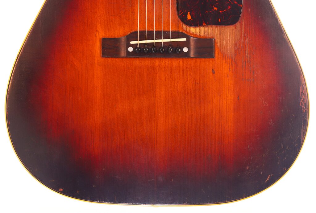Nahaufnahme einer Gibson J-45 1954 Akustische Gitarre mit Sunburst-Lackierung, die den Steg, die Saiten und einen Teil des Schlagbretts zeigt. Sichtbare Kratzer und Gebrauchsspuren auf dem Holz unterstreichen den Vintage-Charakter. Nahaufnahme einer Gibson J-45 1954 Akustische Gitarre mit Sunburst-Lackierung, die den Steg, die Saiten und einen Teil des Schlagbretts zeigt. Sichtbare Kratzer und Gebrauchsspuren auf dem Holz unterstreichen den Vintage-Charakter.