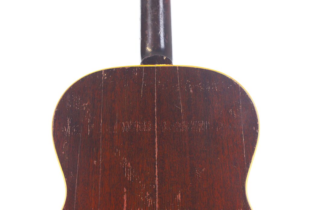 Das Bild zeigt die Rückseite einer abgenutzten Gibson LG-1 1953 Akustik-Gitarre, die eine zentrale Naht und sichtbare Kratzspuren aufweist, fotografiert vor einem weißen Hintergrund. Das Bild zeigt die Rückseite einer abgenutzten Gibson LG-1 1953 Akustik-Gitarre, die eine zentrale Naht und sichtbare Kratzspuren aufweist, fotografiert vor einem weißen Hintergrund.