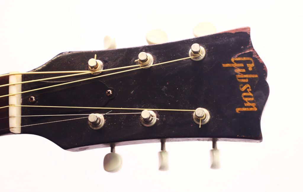 Nahaufnahme der Kopfplatte einer Gibson LG-1 1953 Akustik-Gitarre in Schwarz, mit sechs Stimmwirbeln, Saiten und dem Gibson-Logo in Gold auf weißem Hintergrund. Nahaufnahme der Kopfplatte einer Gibson LG-1 1953 Akustik-Gitarre in Schwarz, mit sechs Stimmwirbeln, Saiten und dem Gibson-Logo in Gold auf weißem Hintergrund.