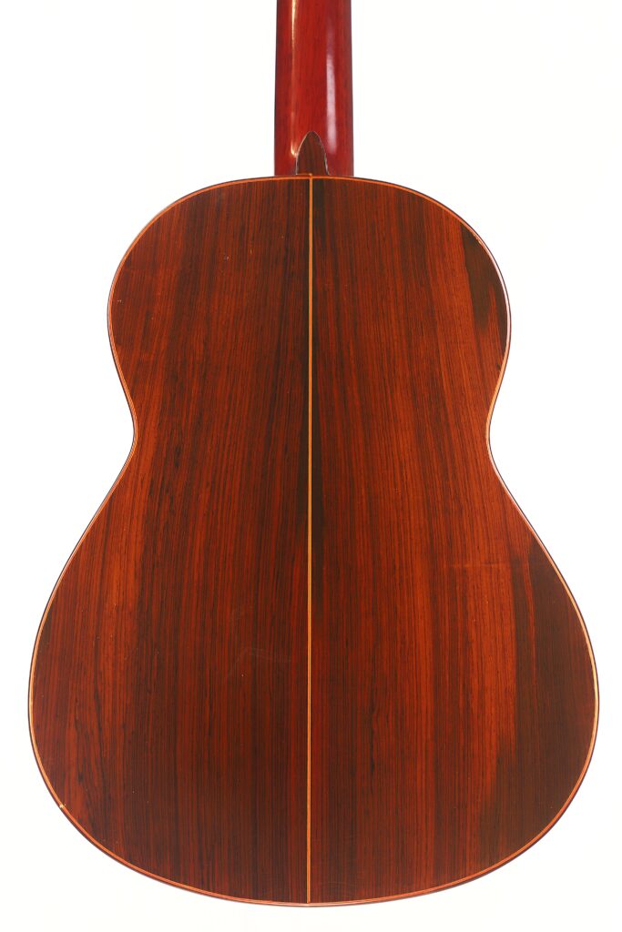 Die Abbildung zeigt die Rückseite einer Faustino Conde Negra Flamenco-Gitarre von Hermanos Conde aus dem Jahr 1971. Sie zeichnet sich durch eine polierte rotbraune Holzoberfläche, geschwungene Kanten, eine glänzende Oberfläche und eine dünne vertikale Naht aus, die in der Mitte verläuft. Die Abbildung zeigt die Rückseite einer Faustino Conde Negra Flamenco-Gitarre von Hermanos Conde aus dem Jahr 1971. Sie zeichnet sich durch eine polierte rotbraune Holzoberfläche, geschwungene Kanten, eine glänzende Oberfläche und eine dünne vertikale Naht aus, die in der Mitte verläuft.
