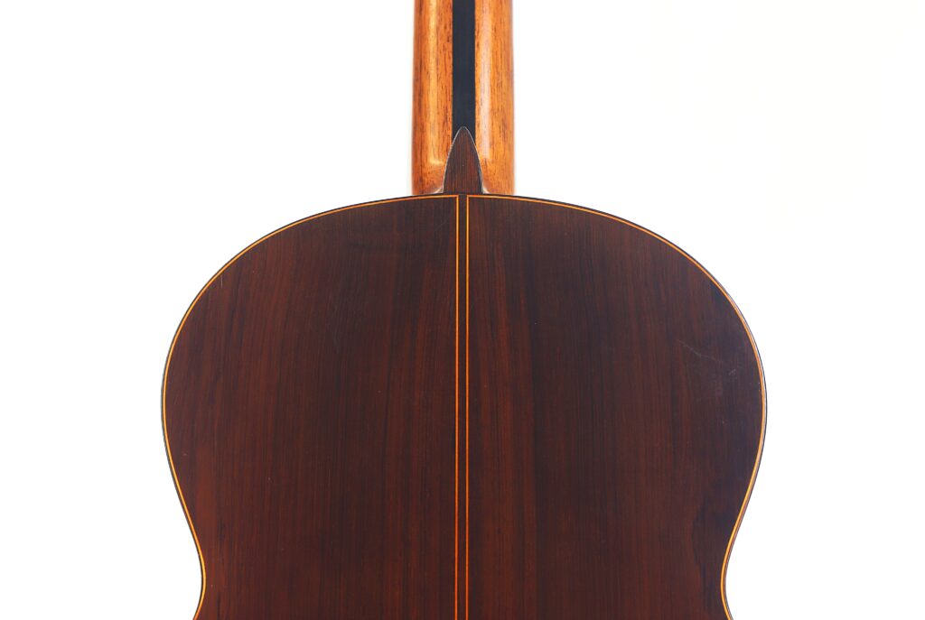 Das Bild zeigt die Rückseite einer Jose Ramirez 1a 1959 PB Klassische Gitarre, die eine dunkle Holzoberfläche mit einer dünnen vertikalen Einlage in der Mitte vor einem schlichten weißen Hintergrund aufweist. Das Bild zeigt die Rückseite einer Jose Ramirez 1a 1959 PB Klassische Gitarre, die eine dunkle Holzoberfläche mit einer dünnen vertikalen Einlage in der Mitte vor einem schlichten weißen Hintergrund aufweist.