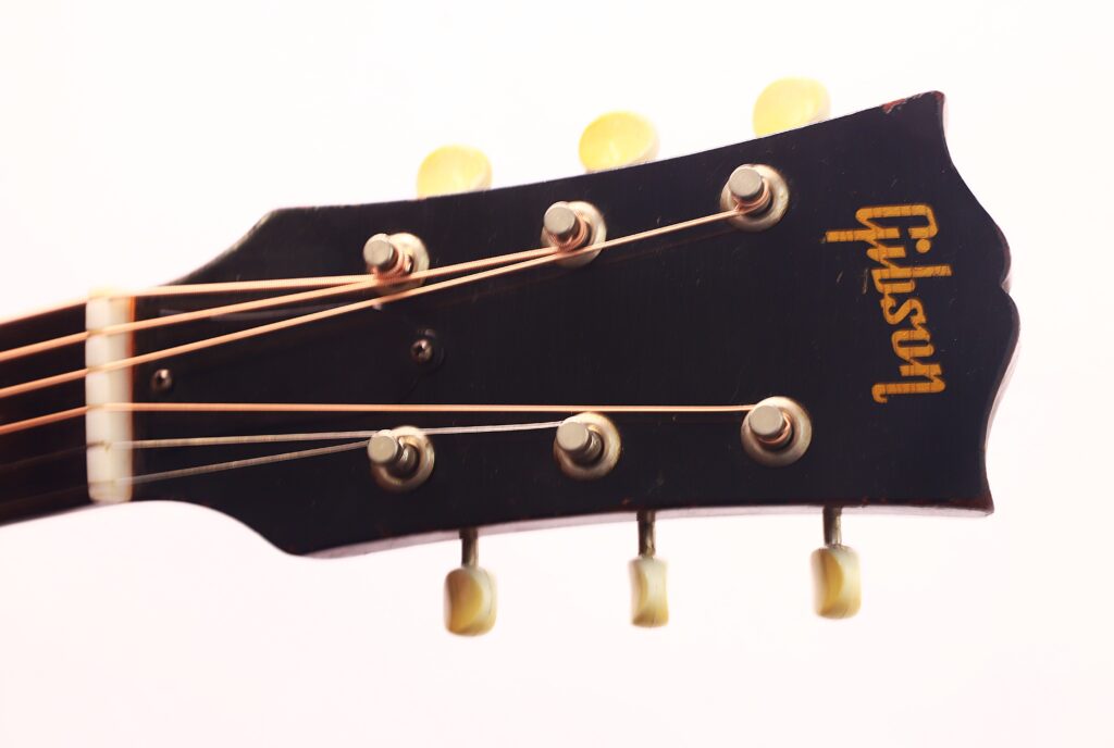 Nahaufnahme einer schwarzen Gibson J-45 1954 Akustische Gitarre mit Kopfplatte, sechs Stimmwirbeln und sichtbaren Saiten vor einem hellen Hintergrund. Das goldene Gibson-Logo ist auf der Kopfplatte aufgedruckt. Nahaufnahme einer schwarzen Gibson J-45 1954 Akustische Gitarre mit Kopfplatte, sechs Stimmwirbeln und sichtbaren Saiten vor einem hellen Hintergrund. Das goldene Gibson-Logo ist auf der Kopfplatte aufgedruckt.