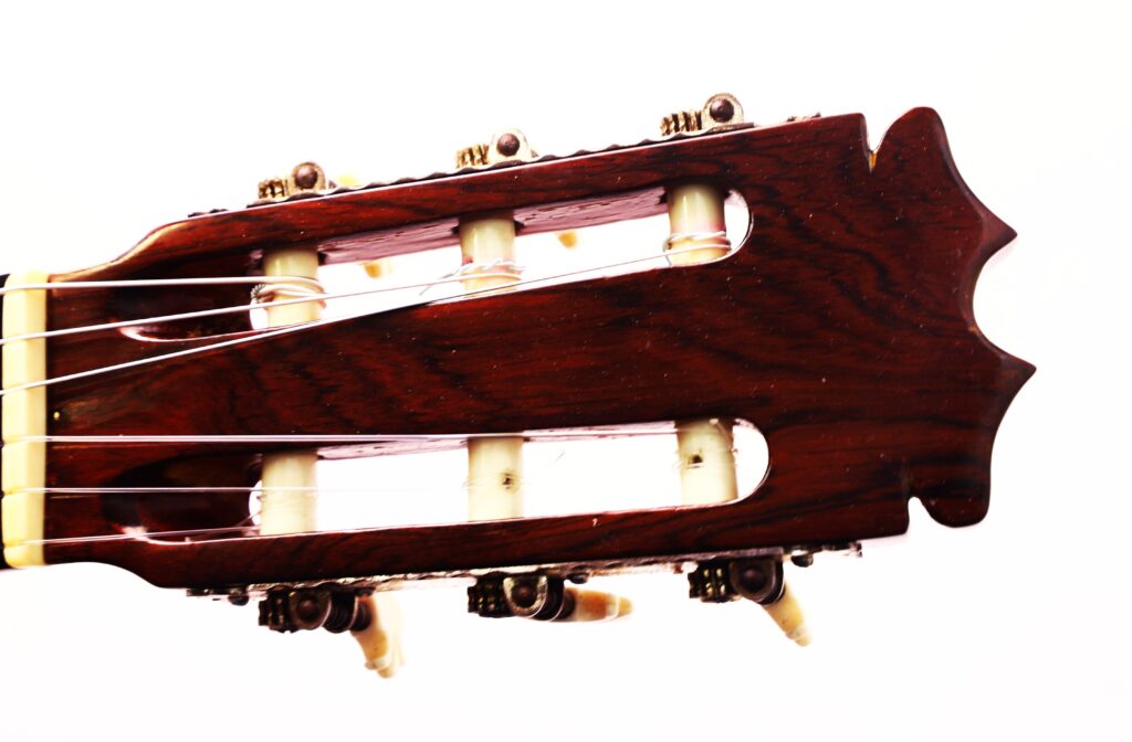 Nahaufnahme der Kopfplatte einer Faustino Conde Negra Flamenco-Gitarre von Hermanos Conde aus dem Jahr 1971 mit hölzernen Mechaniken, cremefarbenen Stimmwirbeln und Gitarrensaiten vor weißem Hintergrund. Nahaufnahme der Kopfplatte einer Faustino Conde Negra Flamenco-Gitarre von Hermanos Conde aus dem Jahr 1971 mit hölzernen Mechaniken, cremefarbenen Stimmwirbeln und Gitarrensaiten vor weißem Hintergrund.