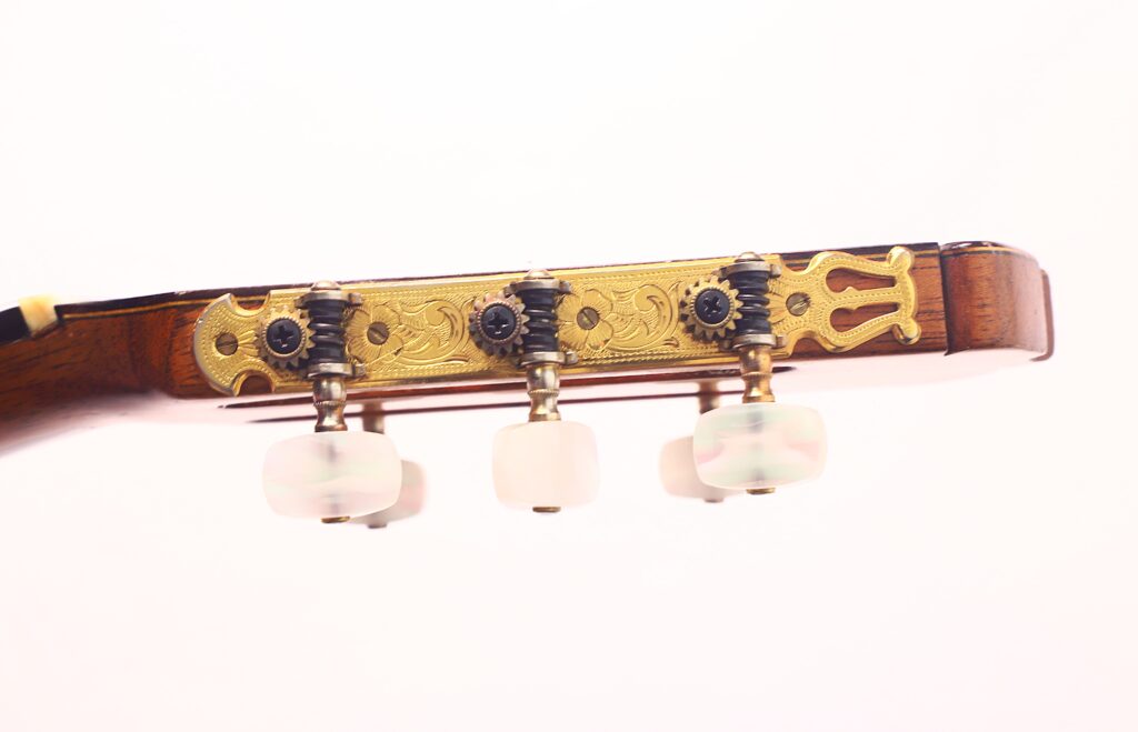 Nahaufnahme der Kopfplatte der Jose Ramirez 1a 1959 PB Klassische Gitarre mit drei weißen Stimmwirbeln auf verschnörkelten goldfarbenen Metallmechaniken vor einem schlichten weißen Hintergrund. Nahaufnahme der Kopfplatte der Jose Ramirez 1a 1959 PB Klassische Gitarre mit drei weißen Stimmwirbeln auf verschnörkelten goldfarbenen Metallmechaniken vor einem schlichten weißen Hintergrund.