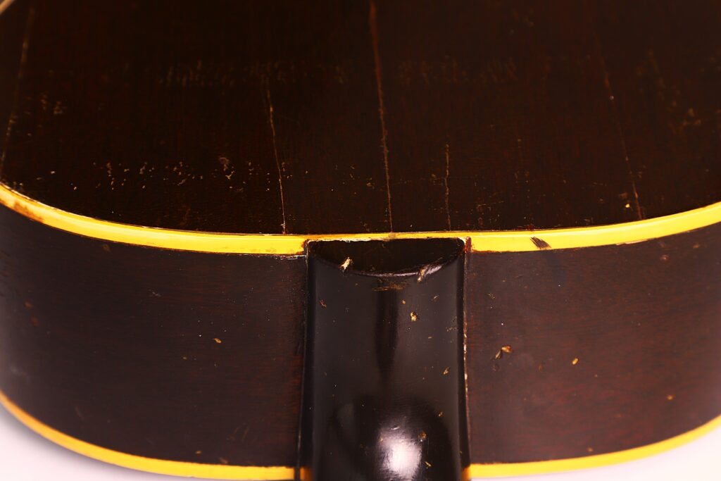 Nahaufnahme der Rückseite und der Seite einer dunkelbraunen Gibson LG-1 1953 Akustik Gitarre mit gelber Zierleiste, wobei die Halsverbindung und sichtbare Oberflächenkratzer hervorgehoben werden. Nahaufnahme der Rückseite und der Seite einer dunkelbraunen Gibson LG-1 1953 Akustik Gitarre mit gelber Zierleiste, wobei die Halsverbindung und sichtbare Oberflächenkratzer hervorgehoben werden.