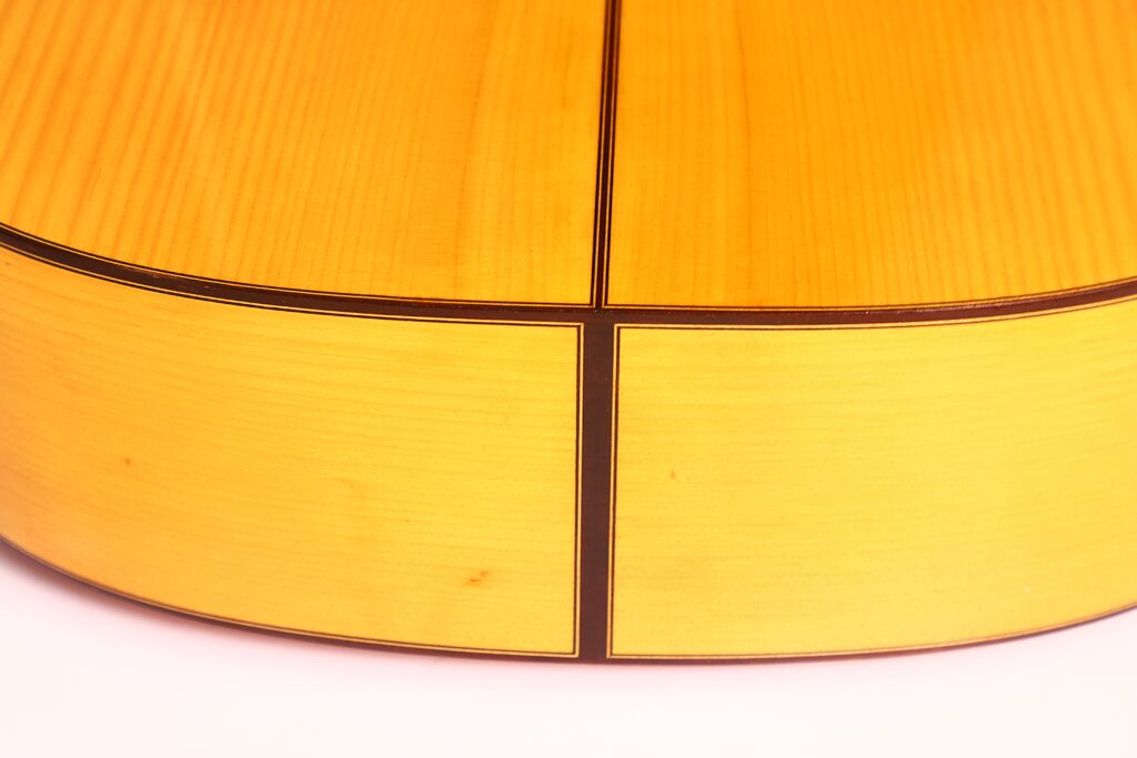 Nahaufnahme der unteren Rückenkante einer Flamenco-Gitarre von Marcelino López Nieto aus dem Jahr 1959. Sie zeigt helles Holz mit dunklen Einlegearbeiten, die an den Verbindungsnähten ein Kreuzmuster bilden. Nahaufnahme der unteren Rückenkante einer Flamenco-Gitarre von Marcelino López Nieto aus dem Jahr 1959. Sie zeigt helles Holz mit dunklen Einlegearbeiten, die an den Verbindungsnähten ein Kreuzmuster bilden.
