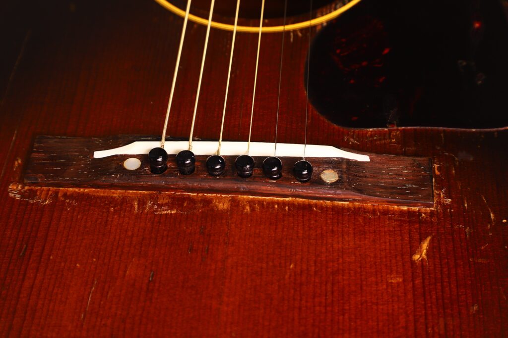 Nahaufnahme der Gibson LG-1 1953 Akustik Gitarre mit sechs Saiten, schwarzen Stegstiften, einem weißen Sattel und einem Holzkorpus mit sichtbarer Maserung und leichten Gebrauchsspuren. Nahaufnahme der Gibson LG-1 1953 Akustik Gitarre mit sechs Saiten, schwarzen Stegstiften, einem weißen Sattel und einem Holzkorpus mit sichtbarer Maserung und leichten Gebrauchsspuren.