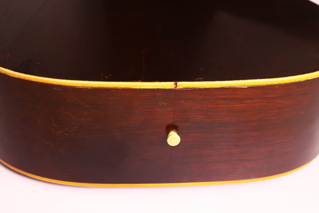 Nahaufnahme der Seite einer Gibson J-45 1954 Akustische Gitarre mit glänzendem dunkelbraunem Finish, gelben Zierleisten und einem einzelnen Metallgurtknopf in der Mitte. Nahaufnahme der Seite einer Gibson J-45 1954 Akustische Gitarre mit glänzendem dunkelbraunem Finish, gelben Zierleisten und einem einzelnen Metallgurtknopf in der Mitte.