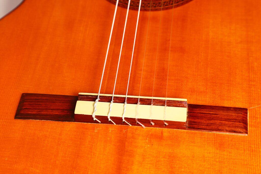 Nahaufnahme der Marcelino López Nieto 1959 Flamenco-Gitarre mit Steg und Saiten, die den Holzkorpus, den weißen Stegsattel und die Nylonsaiten zeigt. Nahaufnahme der Marcelino López Nieto 1959 Flamenco-Gitarre mit Steg und Saiten, die den Holzkorpus, den weißen Stegsattel und die Nylonsaiten zeigt.