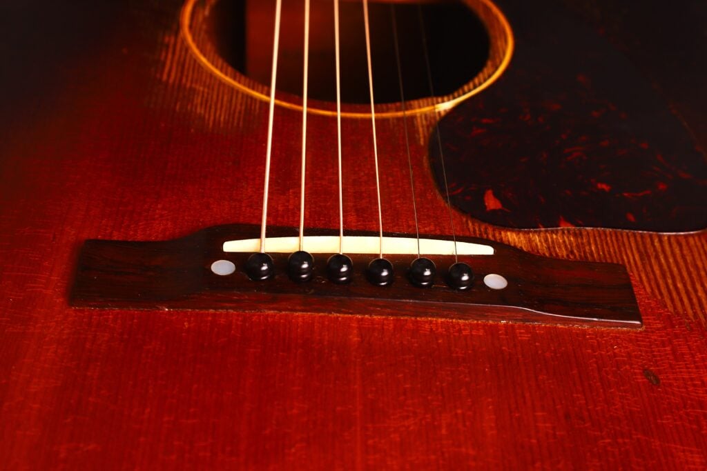 Nahaufnahme des Stegs und der Saiten der Gibson J-45 1954 Akustische Gitarre, mit dunklem Holzsteg, schwarzen Stiften, einem Teil des Schalllochs und einer warmen braunen Lackierung des Korpus. Nahaufnahme des Stegs und der Saiten der Gibson J-45 1954 Akustische Gitarre, mit dunklem Holzsteg, schwarzen Stiften, einem Teil des Schalllochs und einer warmen braunen Lackierung des Korpus.