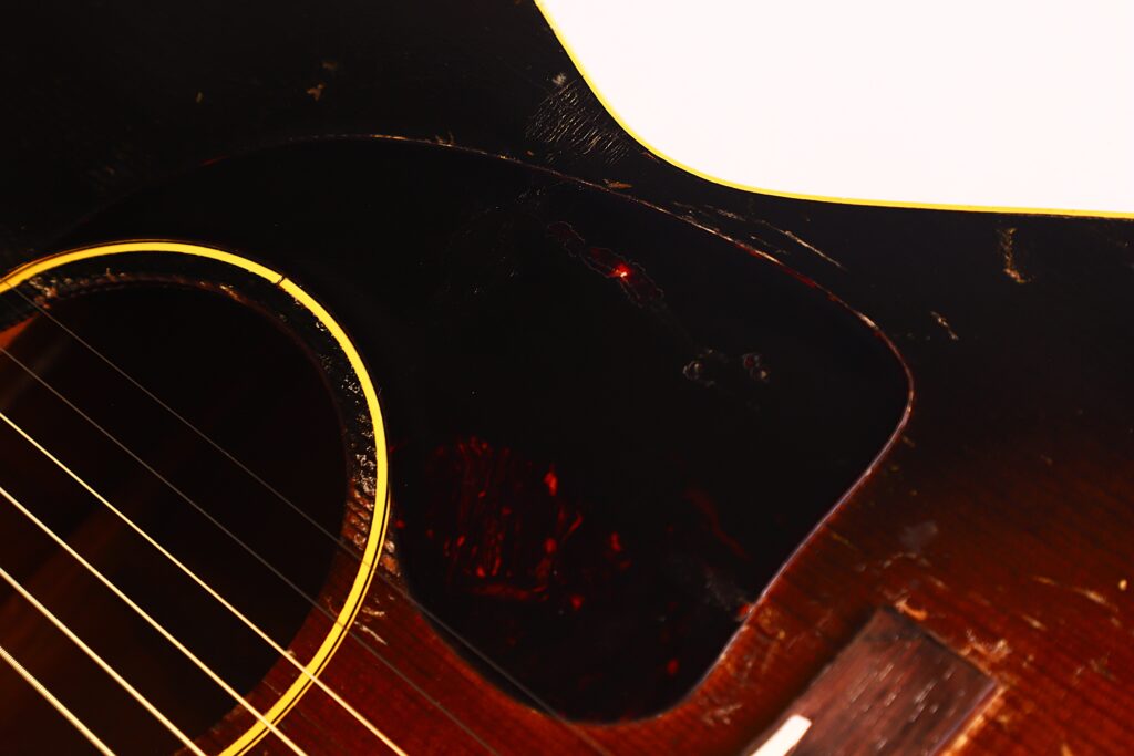 Nahaufnahme der Gibson LG-1 1953 Akustik-Gitarre. Hervorzuheben sind die Sunburst-Lackierung, das Schallloch, die Saiten, das Schlagbrett und kleinere Oberflächenabnutzungen. Nahaufnahme der Gibson LG-1 1953 Akustik-Gitarre. Hervorzuheben sind die Sunburst-Lackierung, das Schallloch, die Saiten, das Schlagbrett und kleinere Oberflächenabnutzungen.