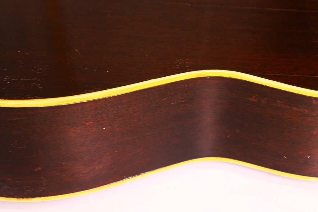 Nahaufnahme des dunkelbraunen Korpus der Gibson LG-1 1953 Akustikgitarre mit gewellten Rändern und einer gelben Zierleiste, die die Kontur umrandet, vor einem hellen Hintergrund. Nahaufnahme des dunkelbraunen Korpus der Gibson LG-1 1953 Akustikgitarre mit gewellten Rändern und einer gelben Zierleiste, die die Kontur umrandet, vor einem hellen Hintergrund.
