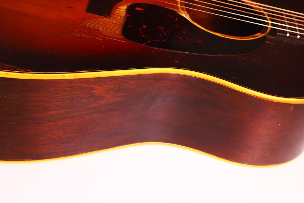 Nahaufnahme einer Gibson J-45 1954 Akustische Gitarre mit dunklem Holzfinish, gelber Zierleiste, sichtbarer Maserung und einem Teil der Saiten und des Schlagbretts vor weißem Hintergrund. Nahaufnahme einer Gibson J-45 1954 Akustische Gitarre mit dunklem Holzfinish, gelber Zierleiste, sichtbarer Maserung und einem Teil der Saiten und des Schlagbretts vor weißem Hintergrund.