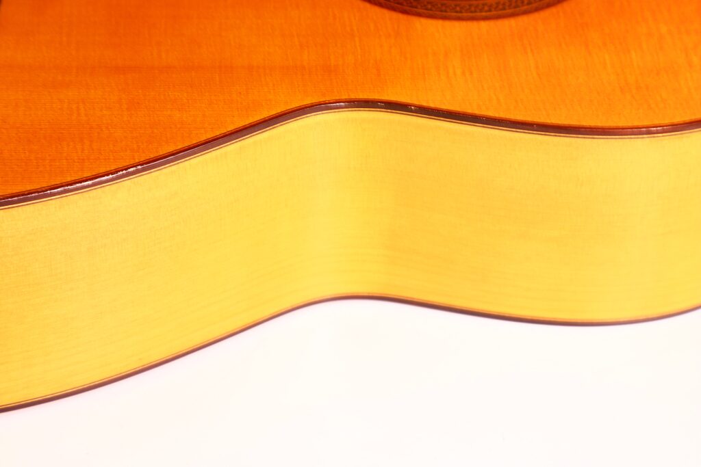 Nahaufnahme der Seite und der Oberkante einer Marcelino López Nieto 1959 Flamenco-Gitarre, die ihre glatte Holzoberfläche und ihre elegant geschwungene Form vor einem schlichten weißen Hintergrund zeigt. Nahaufnahme der Seite und der Oberkante einer Marcelino López Nieto 1959 Flamenco-Gitarre, die ihre glatte Holzoberfläche und ihre elegant geschwungene Form vor einem schlichten weißen Hintergrund zeigt.